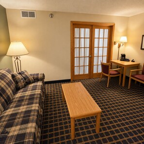 A hotel room with a plaid sofa, a wooden table, a chair, a desk with a lamp, and a patterned carpet. - Moose Lake Lodge & Suites (Moose Lake)