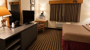 Habitación de hotel con cama, escritorio con computadora y lámpara, televisión, espejo y ventana con cortinas.