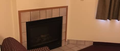 Un salon avec un téléviseur à écran plat posé sur une étagère en bois, au-dessus d’une cheminée recouverte de carreaux. On y trouve un canapé à motifs et un tapis rouge.
