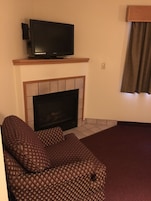 Una sala de estar con una televisión de pantalla plana en un estante de madera sobre una chimenea de baldosas. Hay un sofá estampado y una alfombra roja.