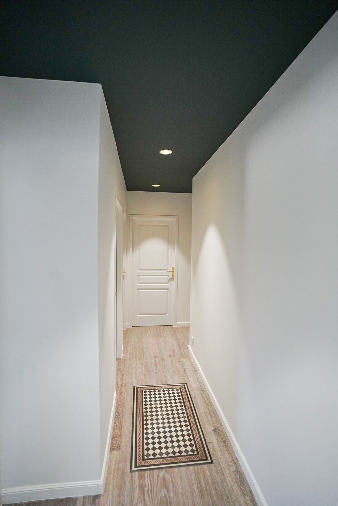 A narrow hallway with a wooden floor, white walls, and a dark ceiling.