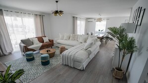A spacious living room with a large sofa set, a coffee table, and a dining area with chairs. There are potted plants and a hanging light fixture.