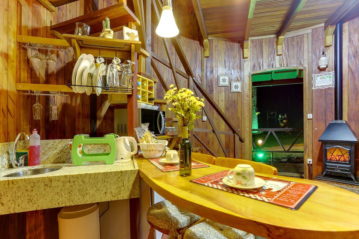 Uma cozinha rústica com paredes de madeira, área de bar com banquetas, lareira e vista para o exterior.