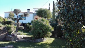 Una propiedad residencial con fachada blanca, una chimenea de piedra y un jardín cuidado con arbustos variados y una palmera.