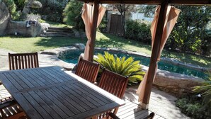 Mesa y sillas de madera bajo una pérgola con vistas a una piscina y jardín.
