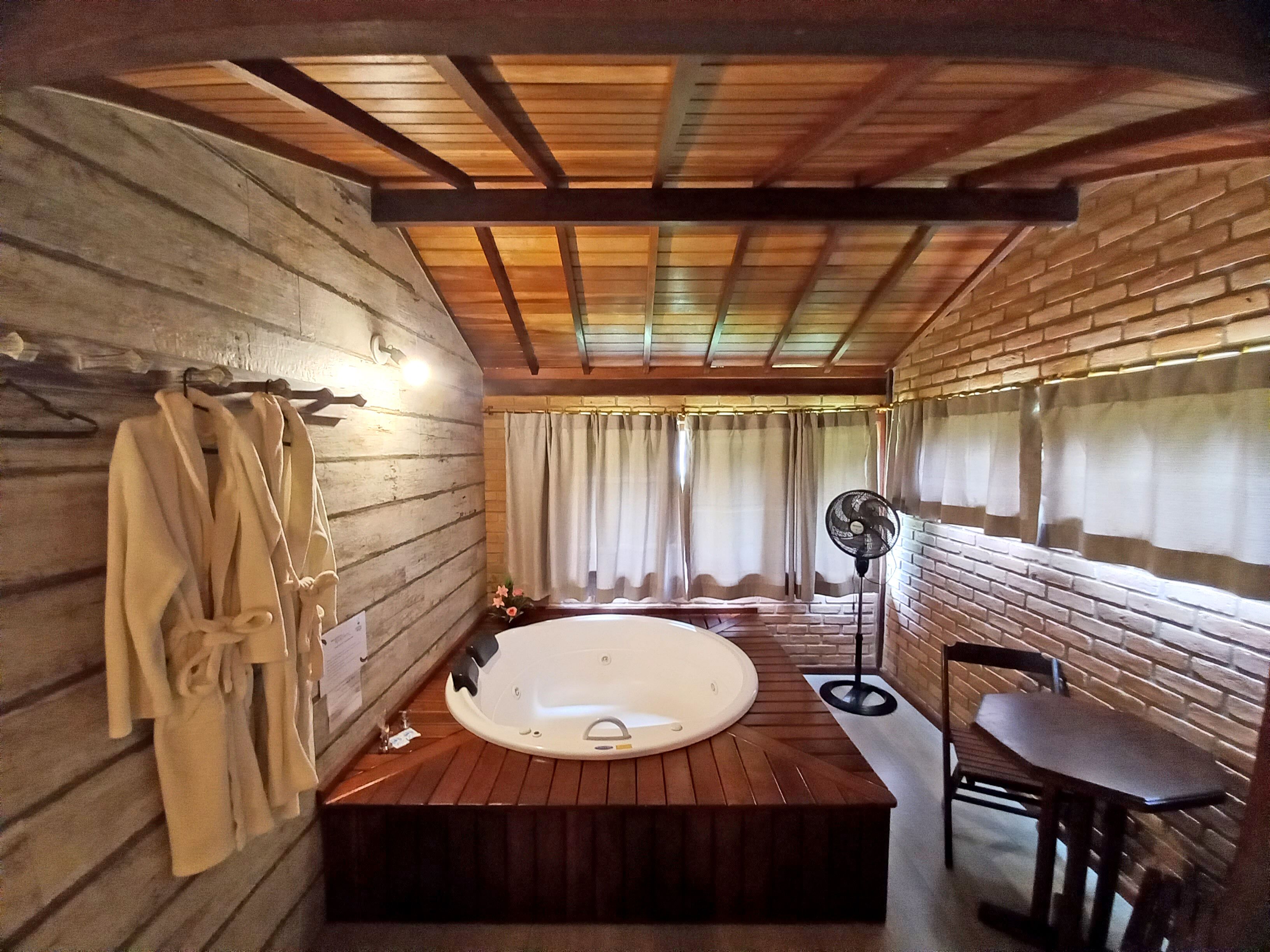 Un baño de cabaña de madera con una bañera de madera, un banco de madera y un ventilador.