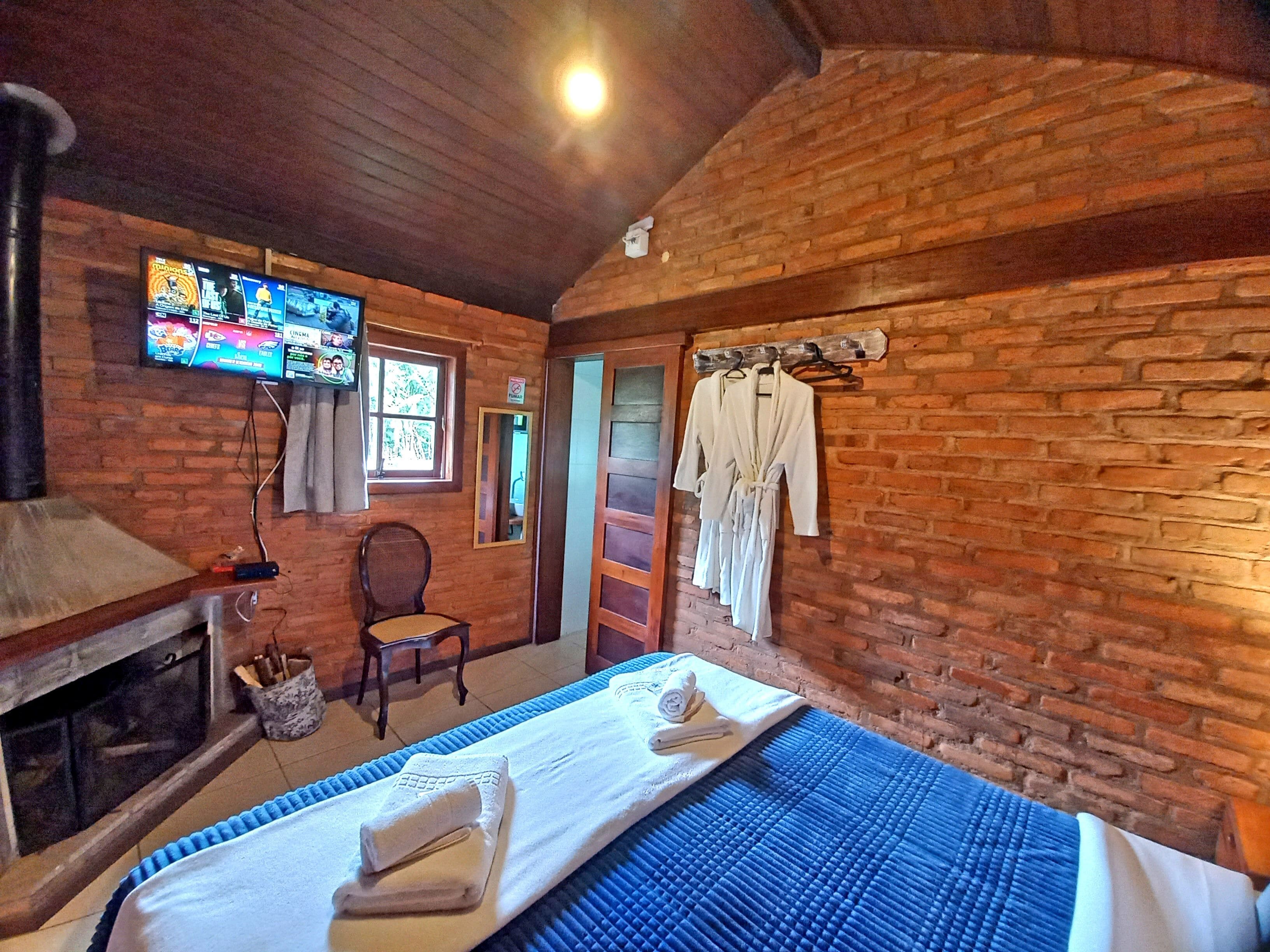 A room with a brick wall, a television mounted on the wall, a bed with a blue mattress, a chair, and a wooden bench.