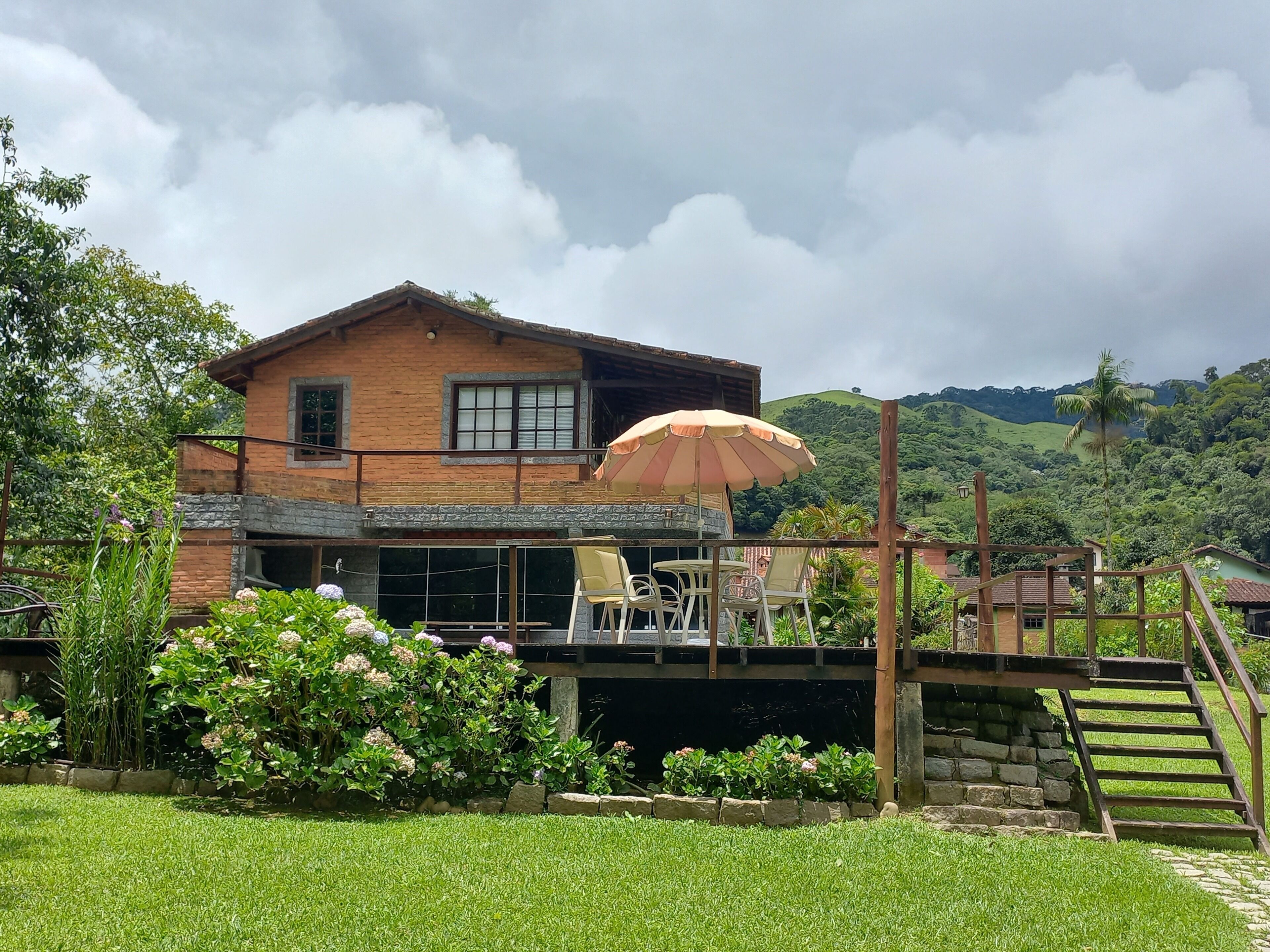 A two-story house with a balcony, a patio with chairs and an umbrella, and a stone staircase leading to a lower level.