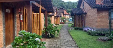 A pathway leading to traditional-style buildings with brick walls and tiled roofs, surrounded by well-maintained gardens and a hilly backdrop.