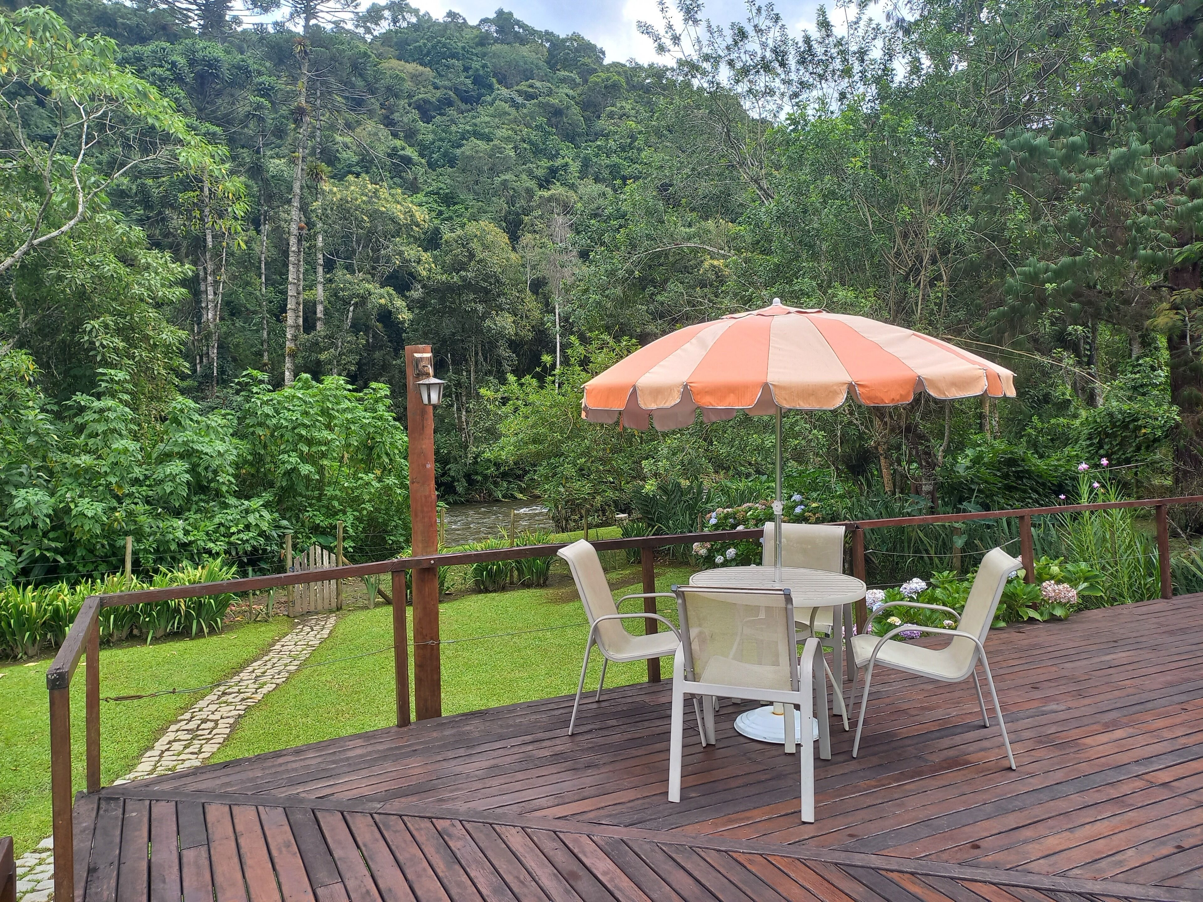 A wooden deck with a table and chairs under an umbrella, surrounded by lush greenery and a river.