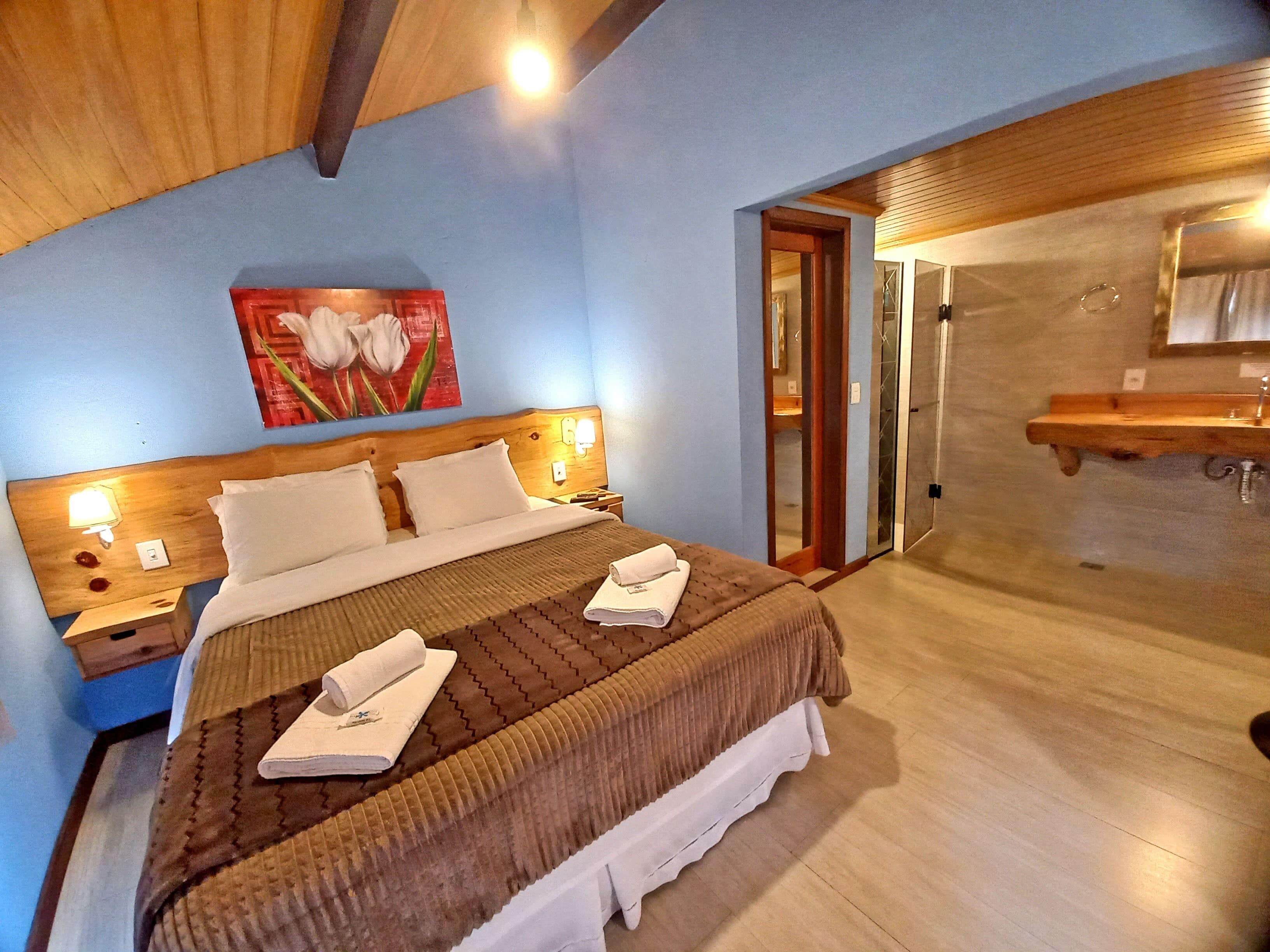 Une chambre d’hôtel avec un grand lit, une tête de lit en bois et un tableau représentant des fleurs. La salle de bain est équipée d’une douche et d’un lavabo.