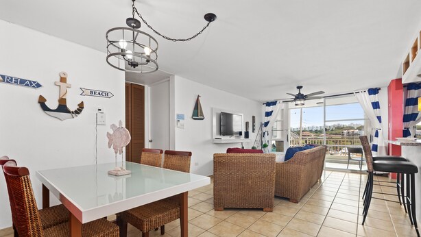 Ein modernes Wohnzimmer mit einem Glastisch, Korbmöbeln, einem Deckenventilator und Blick auf die Stadt durch große Fenster.