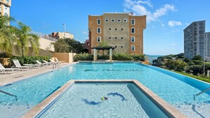 Ein Schwimmbecken mit Rettungsschwimmerstation, Liegestühlen und Blick auf ein mehrstöckiges Gebäude.