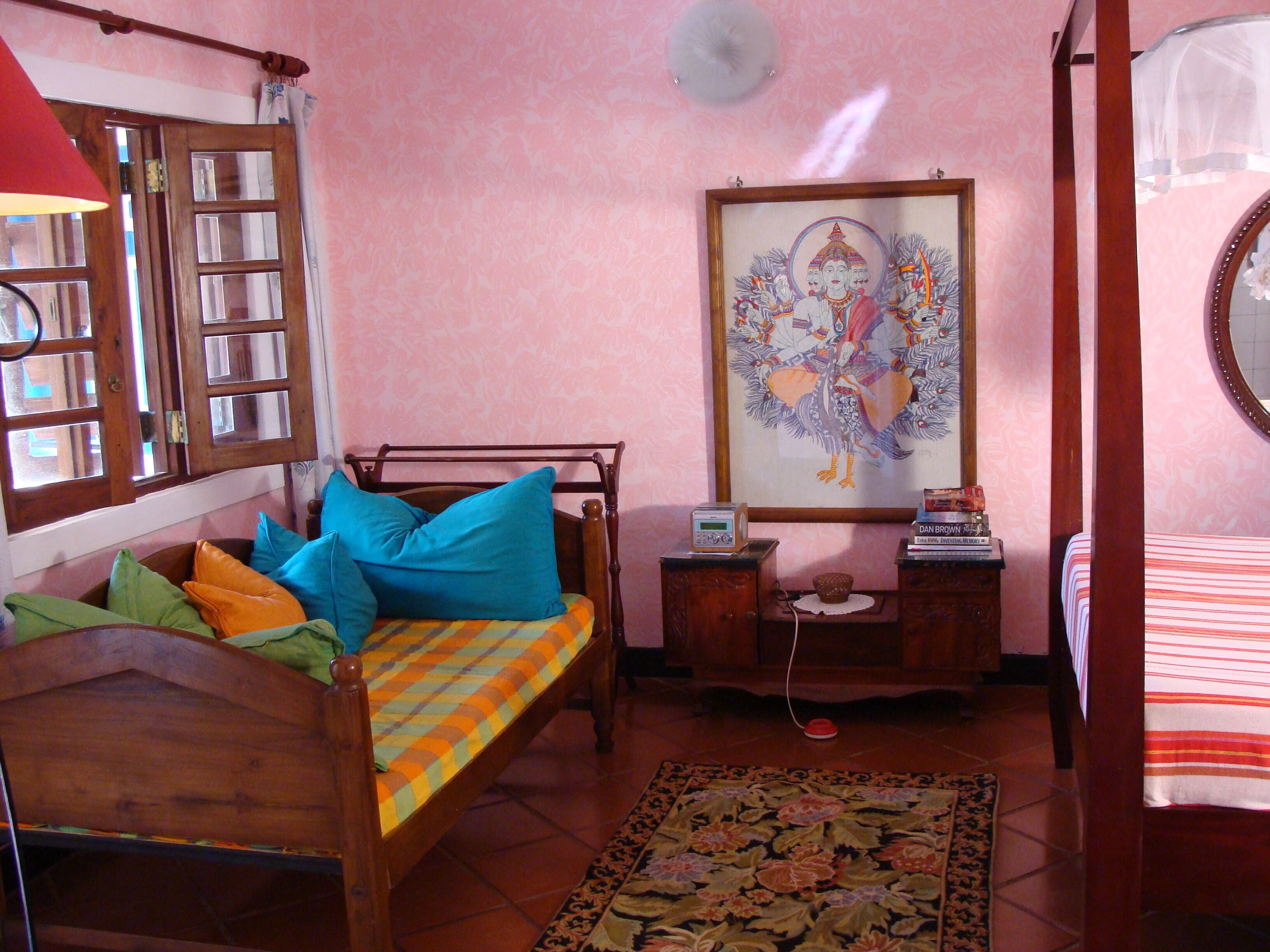 Un dormitorio con cama de madera, almohadas coloridas, una mesita de noche con un radio, un cuadro en la pared y un tapete estampado en el suelo.