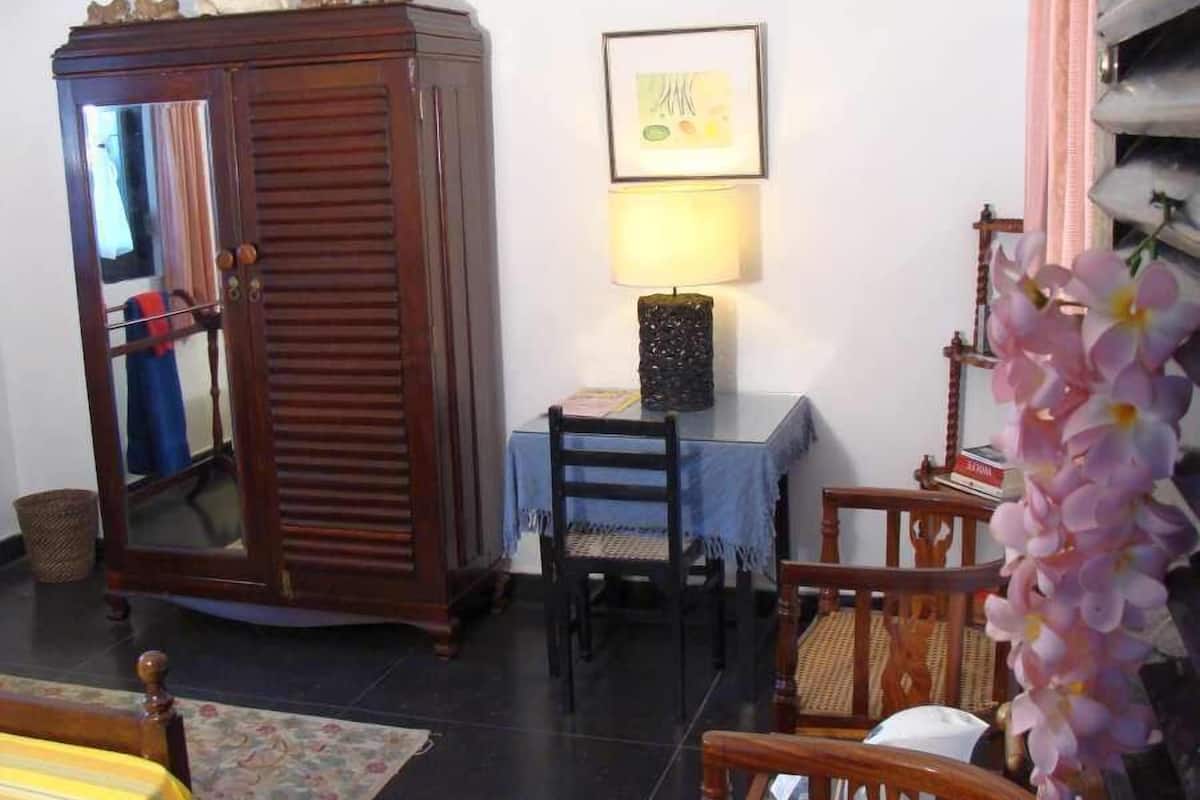 A room with a wooden wardrobe, a table with a lamp, and a chair.