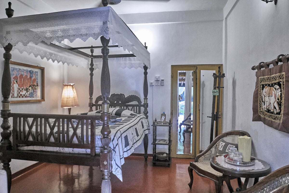 A four-poster bed with a lace canopy, a bedside table with a lamp, a chair with a tray, and a framed picture on the wall.
