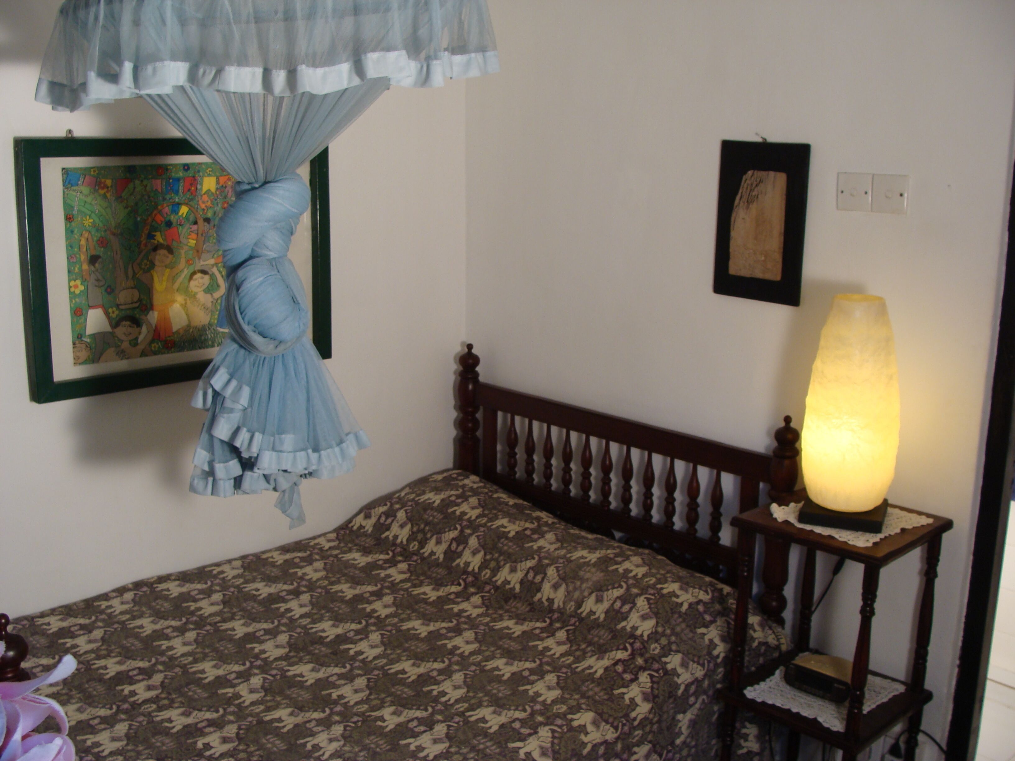 A bedroom with a bed, a nightstand with a lamp, a framed picture on the wall, and a hanging blue fabric.