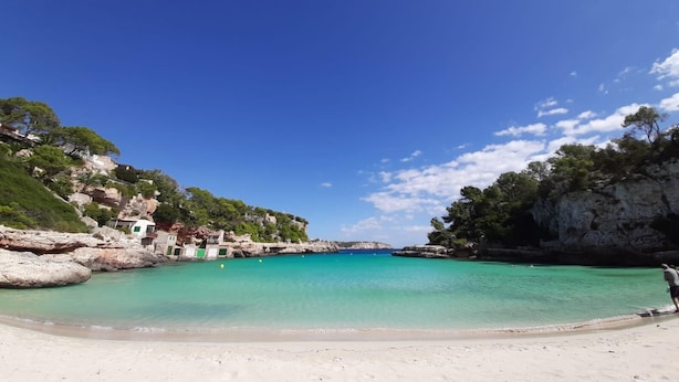 A beach with clear turquoise water, rocky cliffs, and a few buildings on the left. - Casa Sigrunella++with infinity pool (12x4m)++ in Cala Llombards (Cala Llombards)