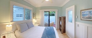 Ein Schlafzimmer mit Bett, Nachttischen, einer Lampe, einem Fenster mit Jalousien, einer Tür zu einem Balkon und einem gerahmten Bild an der Wand.