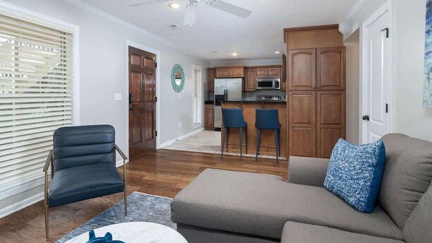 A modern living room with a sofa, a chair, a ceiling fan, and a kitchen area with wooden cabinets and a microwave.