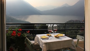 Een tafel met een tafelkleed, twee wijnglazen en borden met eten op een balkon met uitzicht op een meer en bergen.