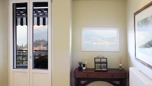 Een kamer met uitzicht op een stad door een raam, een lijst met een foto aan de muur, en een klein tafeltje met decoratieve objecten.