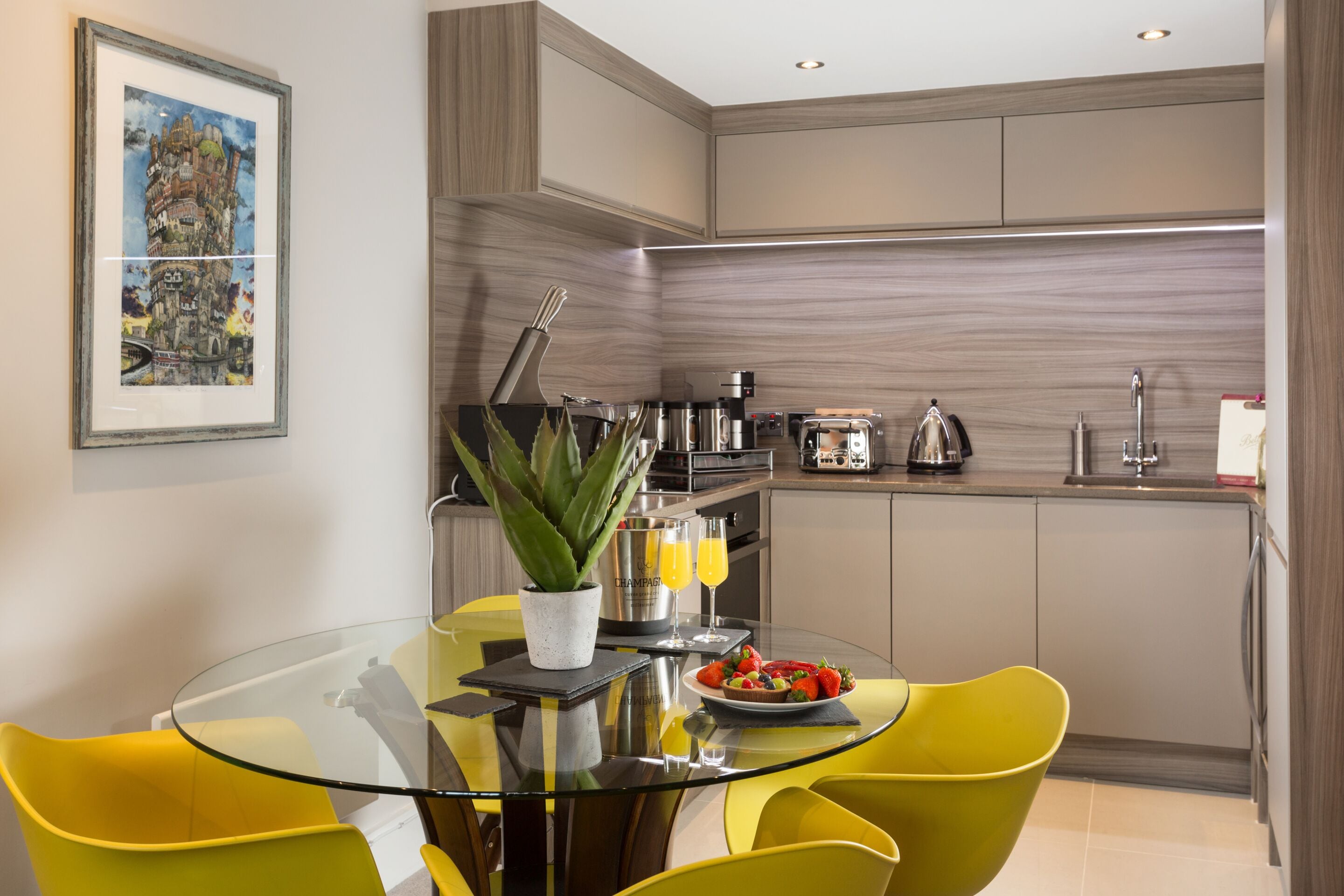A modern kitchen with a glass dining table, yellow chairs, and a potted plant.