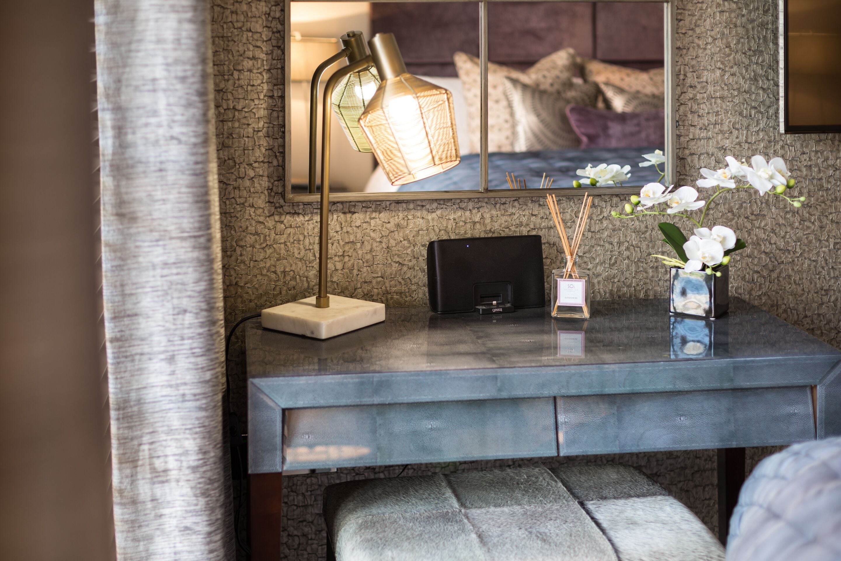A modern room with a desk, a lamp, a vase with flowers, and a mirror reflecting a bed.