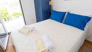 Ein ordentlich bezogenes Bett mit weißer und blauer Bettwäsche, ein kleiner Strauß mit einer gelben Blume und ein blauer Kleiderschrank in einem hellen Zimmer mit Blick auf Grünfläche.