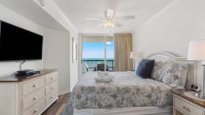 A hotel room with a bed, a TV, a ceiling fan, a balcony with a view of the ocean, and a desk with a lamp. - Ashworth 1809 4 Bedroom Condo by RedAwning (North Myrtle Beach)