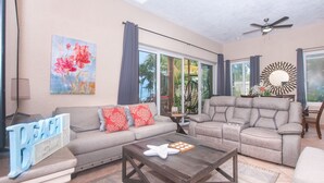 A spacious living room with a grey sofa, a recliner, a coffee table, and a large window offering a view of palm trees. - Coral Sands #6 2 Bedroom Condo by RedAwning (Roatan)