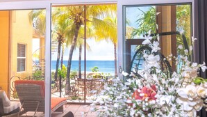 A balcony with wicker chairs, a table, and a view of the ocean and palm trees. - Coral Sands #6 2 Bedroom Condo by RedAwning (Roatan)