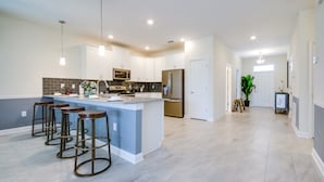 A modern kitchen with a central island, bar stools, stainless steel appliances, and a hallway with a potted plant. - Villa in Champions Gate Resort 2527 (Davenport)