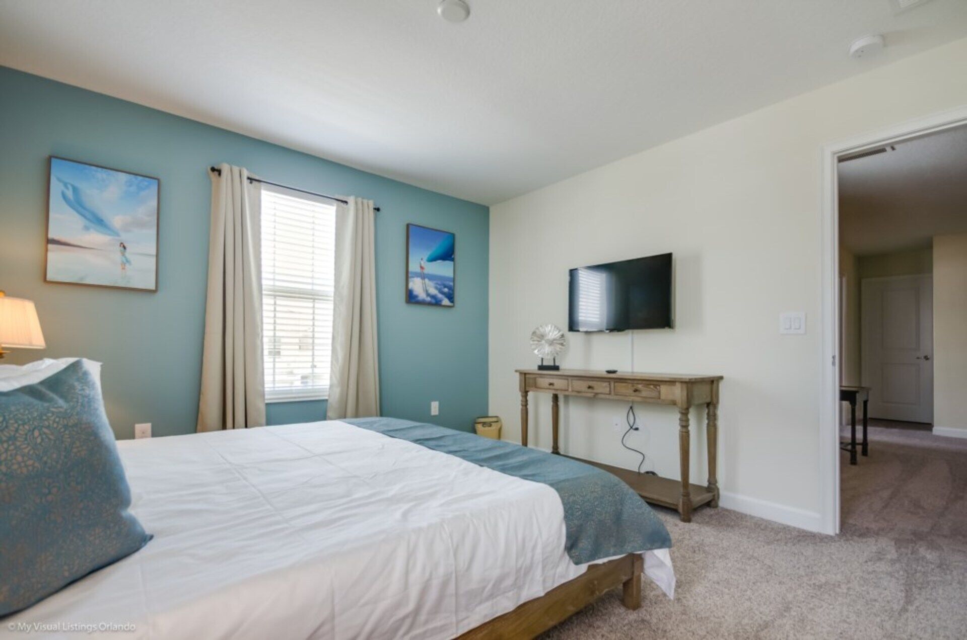 A bedroom with a bed, a desk, a TV, and a door leading to another room.