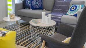 Een moderne woonkamer met een grijze bank, decoratieve kussens, een gele fauteuil en een gestreept vloerkleed. Er staan twee witte ronde tafels met decoratieve objecten op.