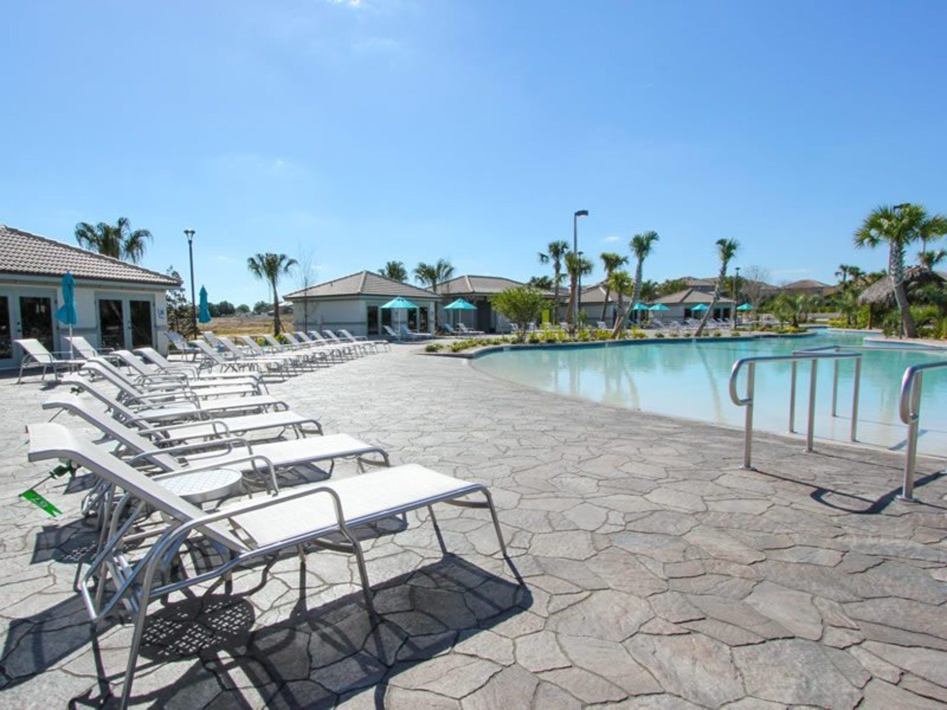 Un espace piscine doté de chaises longues et de parasols, entouré de palmiers et d’immeubles.