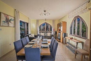 A dining area with a long table set for four, a sofa, and a TV stand. - Private Villa in Pernera 1233 (Paralimni)
