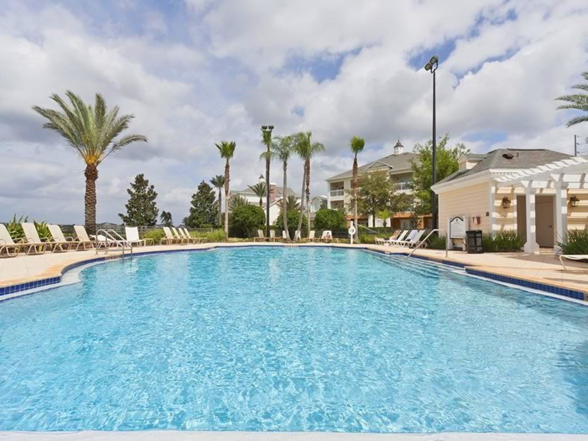 Una piscina azul clara rodeada de sillas de descanso y palmeras, con un edificio al fondo.