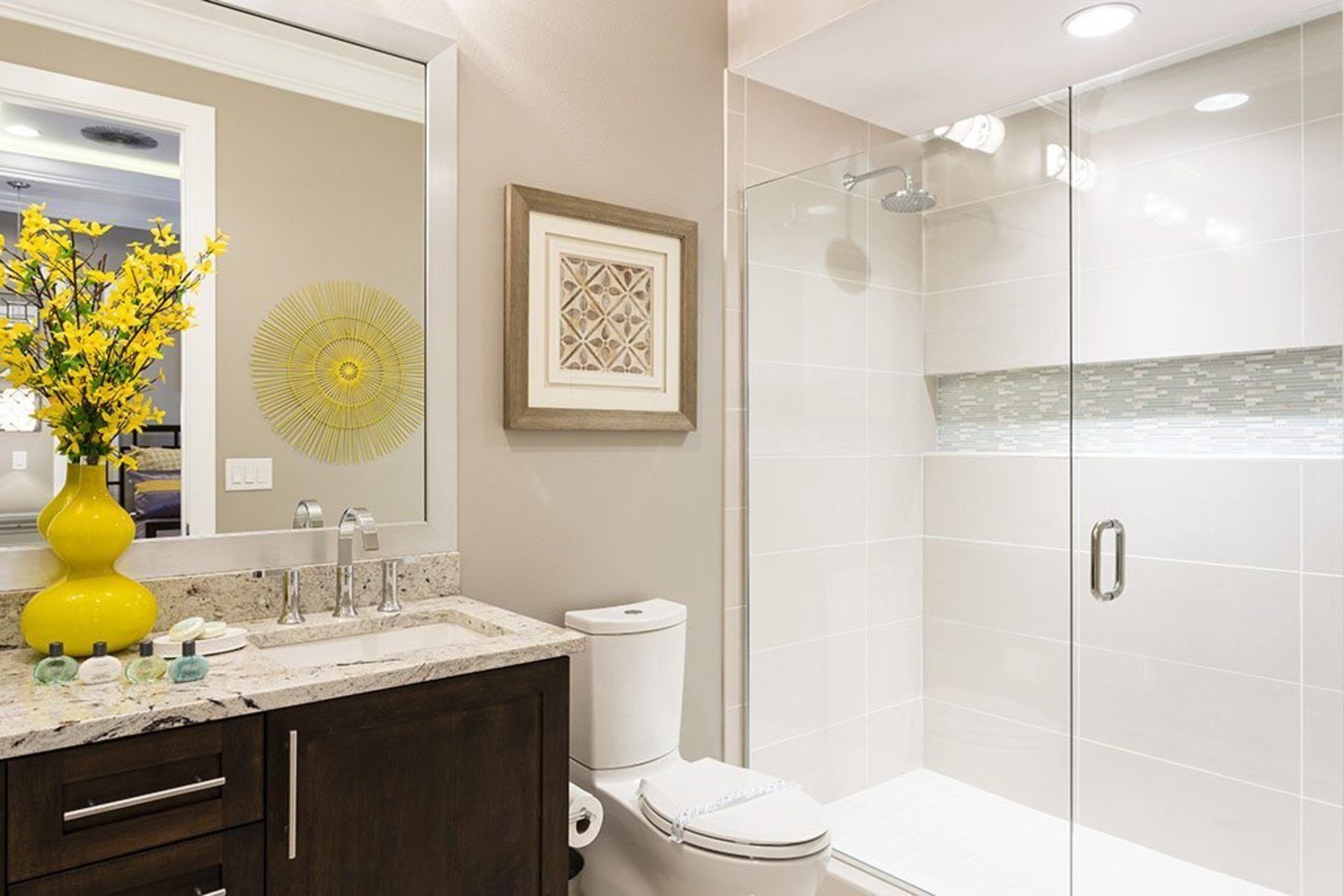 A modern bathroom with a glass shower enclosure, a sink with a marble countertop, a toilet, and a yellow vase with flowers.
