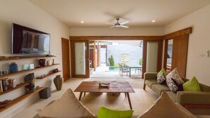 A living room with a TV, shelves with books and decorative items, a coffee table, and a sofa with cushions. There is a ceiling fan and a view of an outdoor area with a pool and greenery. - Private Villa in Seminyak 1150 (Seminyak)