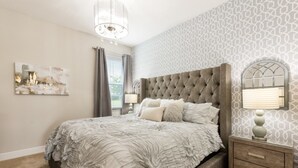 A bedroom with a tufted headboard, a large bed with a ruffled white bedspread, a nightstand with a lamp, and a wall-mounted mirror. - Villa in Encore Resort at Reunion 3009 (Kissimmee)