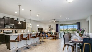 A modern kitchen with a central island, bar stools, and a dining area with a long table set for six. - Villa in Windsor at Westside Resort 3090 (Kissimmee)