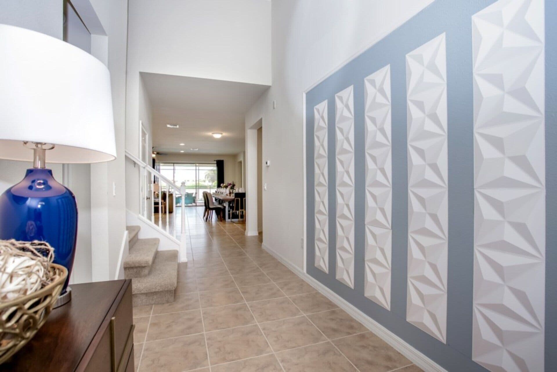 A hallway with a staircase, a dining area in the background, and a wall with geometric patterns.