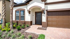A two-story house with a brown garage door, a black front door, and a small porch area with a brick pathway. - Villa in Windsor at Westside Resort 3090 (Kissimmee)