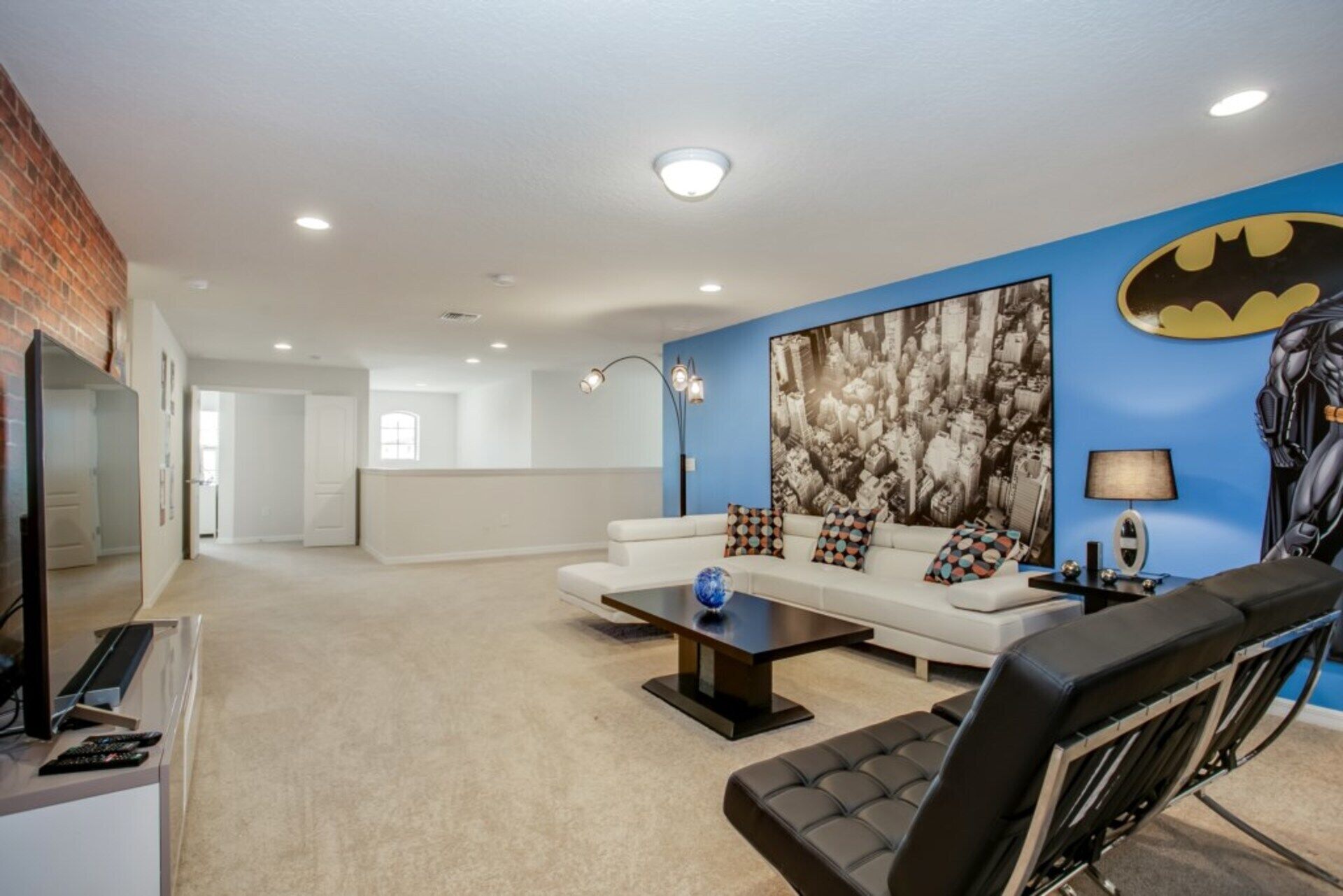 A modern living room with a white sofa, a black leather chair, and a large Batman-themed wall mural.