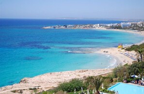 A coastal view with a sandy beach, a clear blue sea, and a resort area with buildings and a swimming pool.