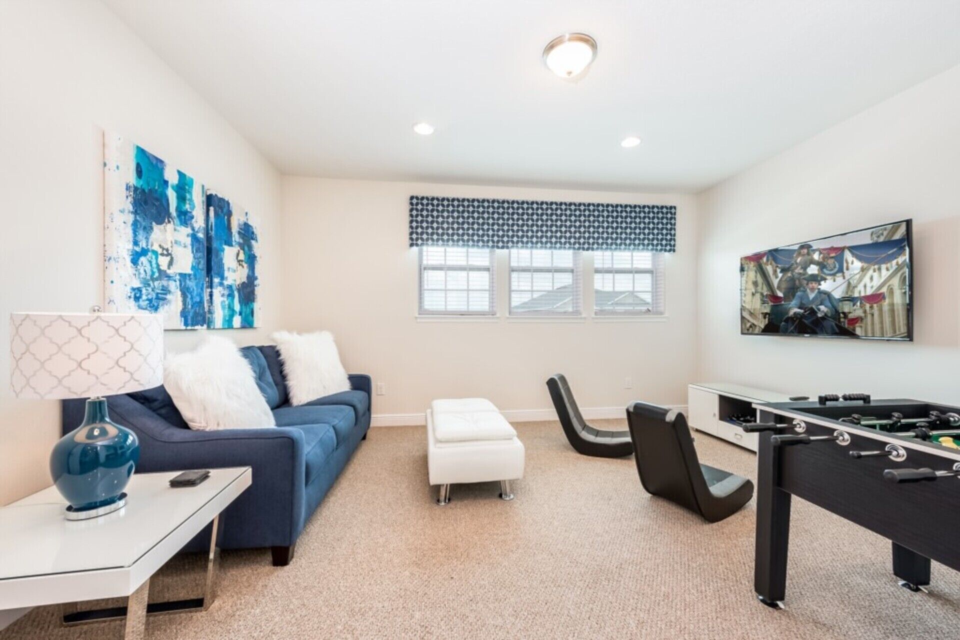 A modern living room with a blue sofa, a white ottoman, a coffee table, a lamp, a foosball table, and a large window with a patterned valance.