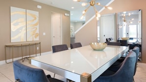 A modern dining area with a large glass table, blue upholstered chairs, and a central light fixture. - Villa in Encore Resort at Reunion 2986 (Kissimmee)