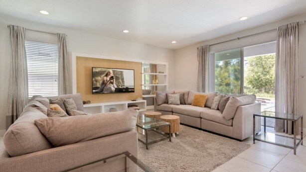 A modern living room with a large flat-screen TV, a sectional sofa, a glass coffee table, and a view of the outdoors through large windows. - Villa in Encore Resort at Reunion 2986 (Kissimmee)