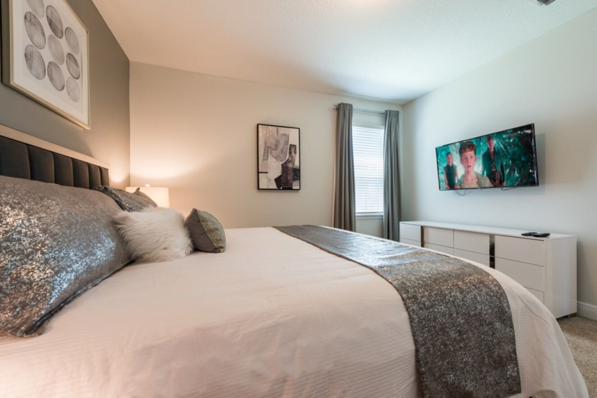 Un dormitorio con una cama grande, un televisor de pantalla plana montado en la pared, una mesita de noche y una ventana con cortinas.